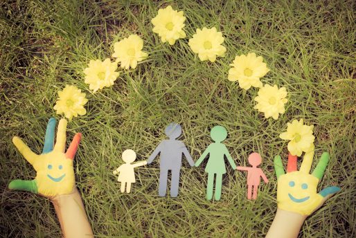 mains d'un enfant, peinturé de manière coloré, placé dans le gazon et entouré de fleurs avec, dans le centre, un papier qui a été découpé de sorte à créer une famille.